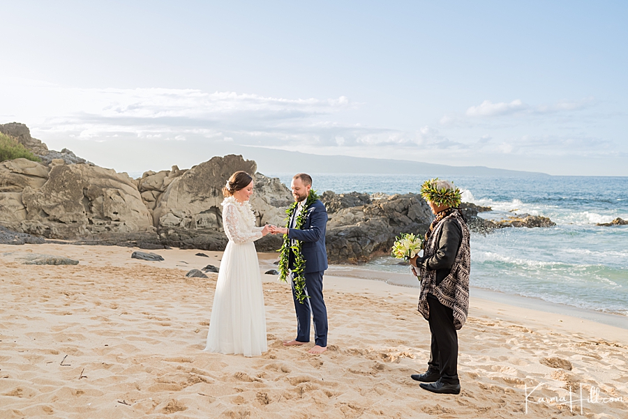 bride wearing long sleeved lace dress and groom in navy blue exchange rings on the beach with hawaiian minister looking on 