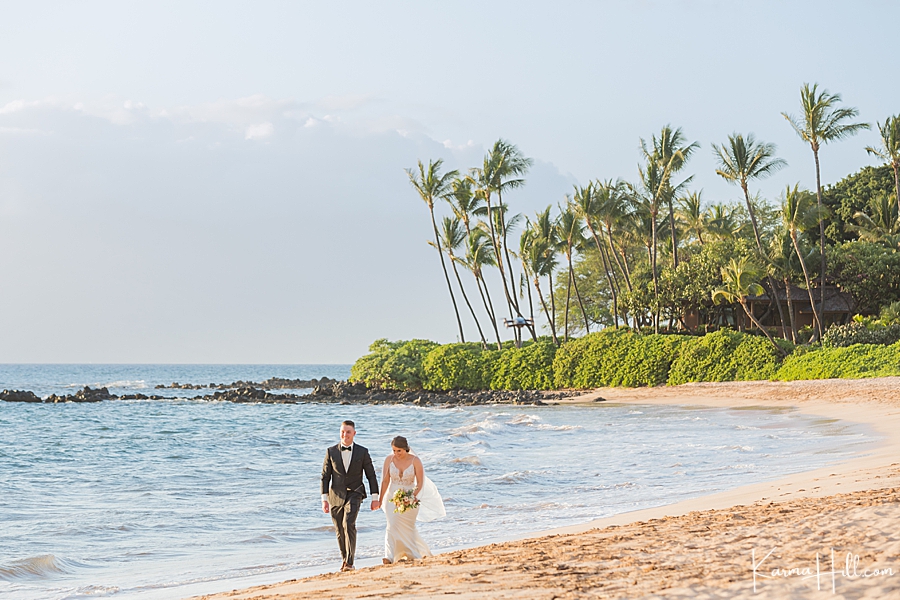 bride and groom walk together holding hands on maui beach with palm trees in the background 