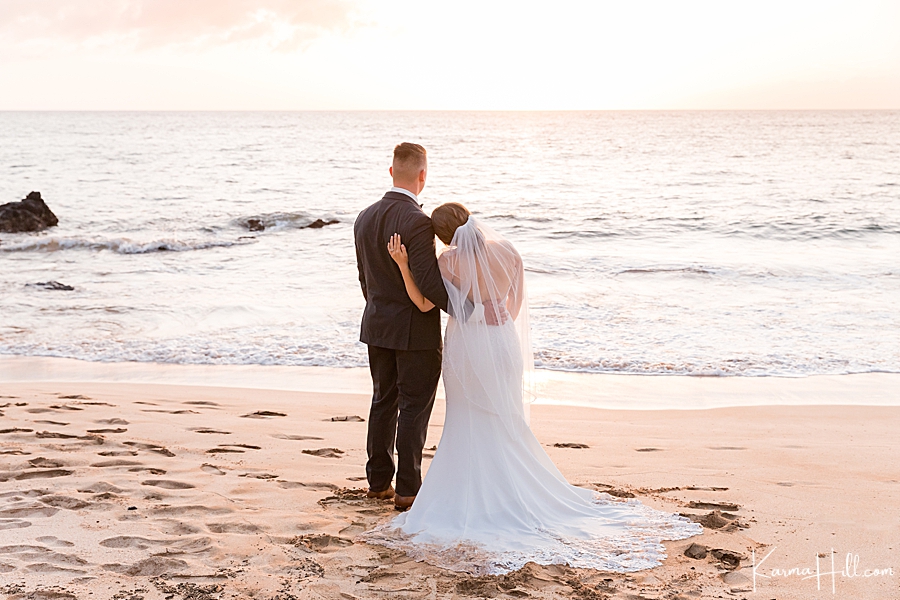 bride places her head on groom's shoulders as they watch a sunset in hawaii 