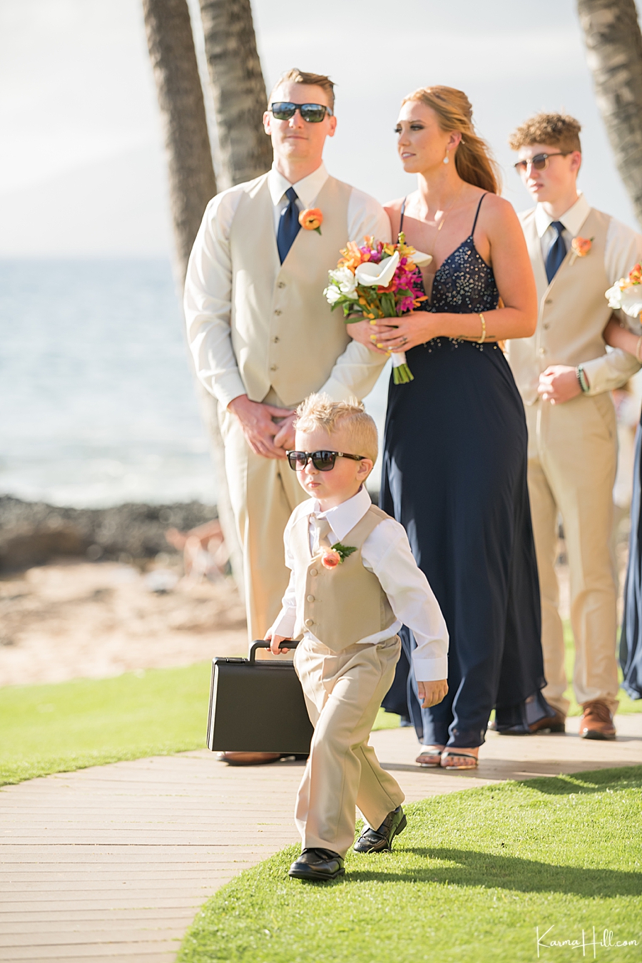 cute ring bearer walks down the aisle for a beach wedding wearing sunglasses and holding a briefcase like a spy 