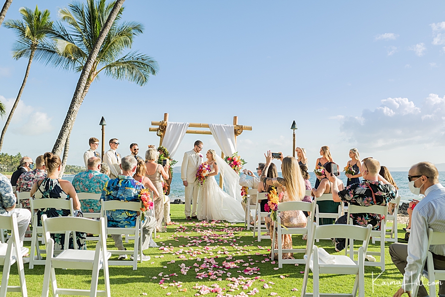 bride and groom stand beneath a white fabric arch with guests watching on and palm trees and ocean in the background 