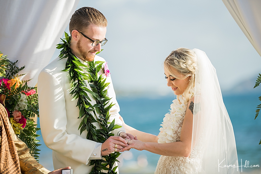 groom puts the ring on bride's finger in front of the ocean 