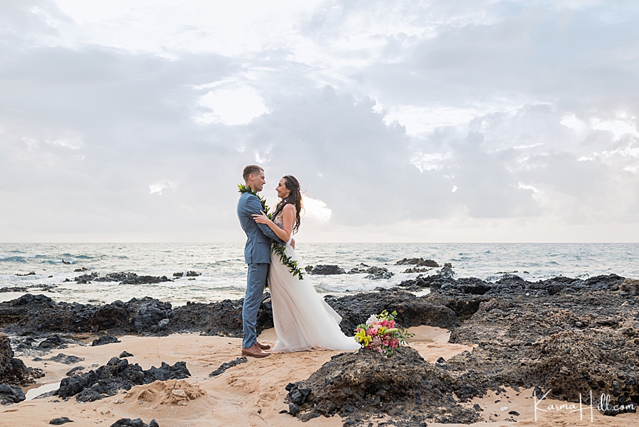 bride and groom hold each other on maui beach with lava rocks and ocean in the background 