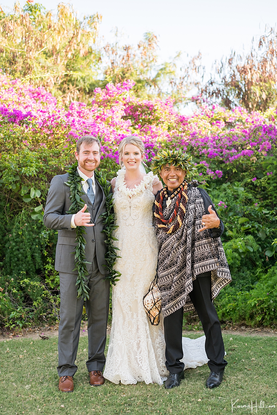 couple poses with hawaiian reverend in front of fuchsia flowers 
