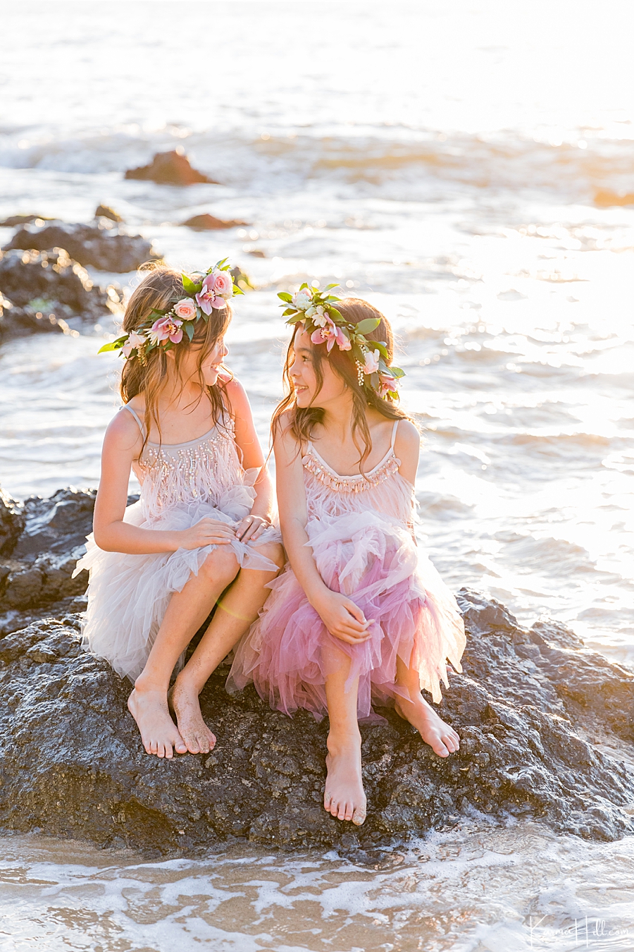 adorable little girls in fluffy dresses and flower crowns sit on a rock in a beach in hawaii 
