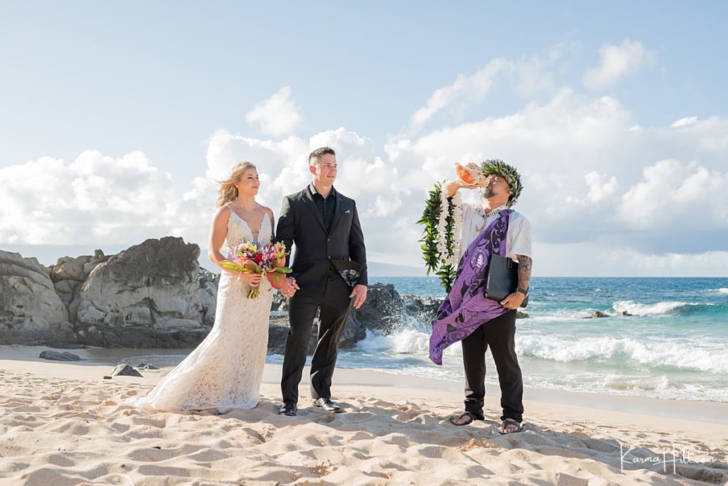 bride and groom stand on ironwoods beach as a hawaiian minister blows the conch shell 