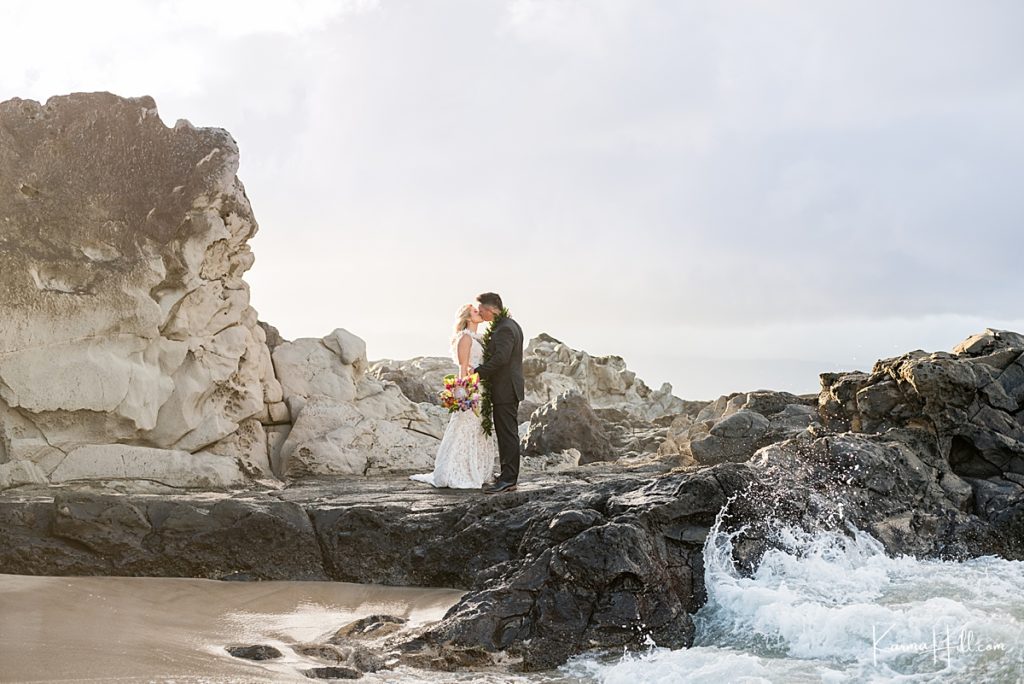 newly wed husband and wife stand on rocky embankment and kiss with crashing waves 