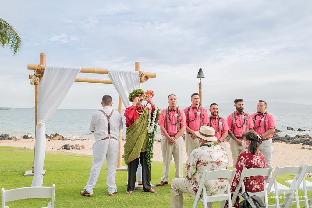 hawaiian minister blows the conch shell as groom waits with groomsmen in pink shirts and trousers 