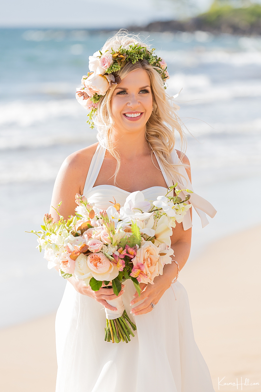bride with blonde hair and flower crown carries an orchid lei on the beach 