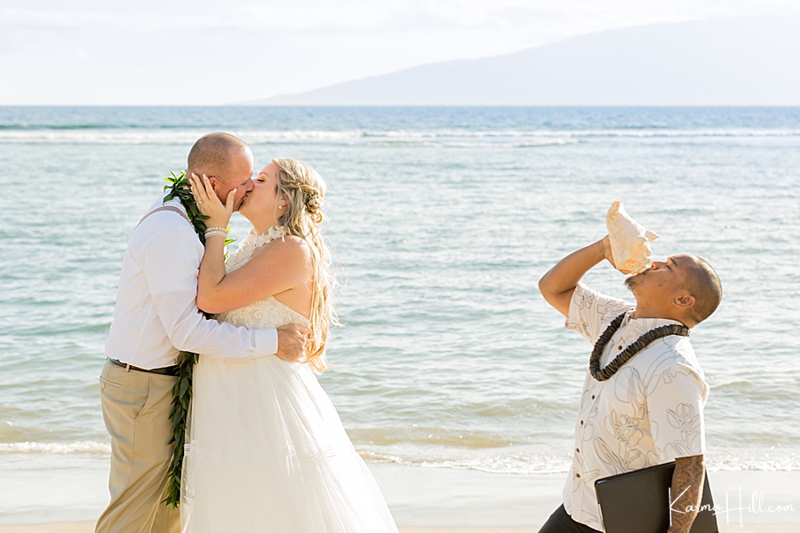hawaiian minister blows the conch shell as couple share their first kiss during their hawaii wedding on the beach 